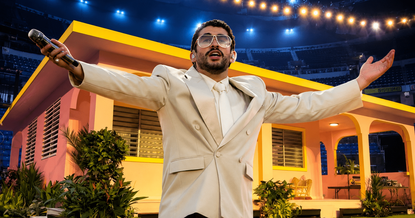 Cantante con traje beige y micrófono posa con los brazos abiertos frente a una casita color rosa y amarillo en un escenario iluminado, con plantas alrededor y luces de concierto al fondo.
