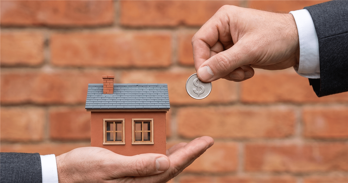 Manos intercambiando una casa en miniatura y una moneda frente a un muro de ladrillo